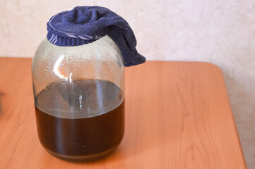 Glass jar with dark liquid covered by a cloth, placed on a wooden table. Concept of homemade...