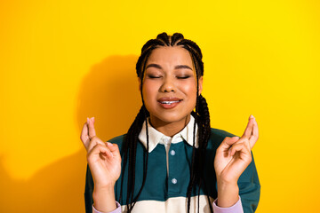 Portrait of a young woman with braided hair on yellow background expressing hope with crossed...