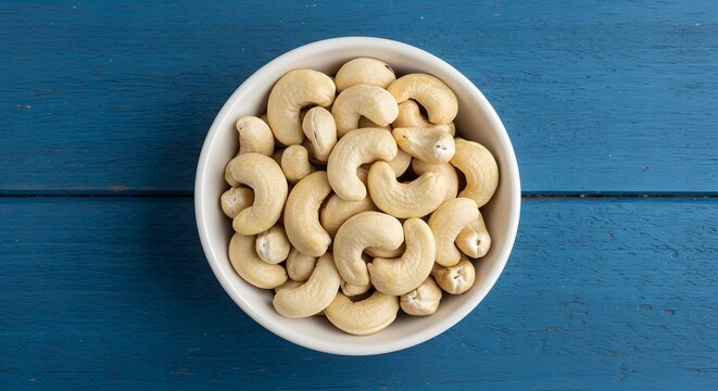 A top view of a white bowl filled with cashew nuts on a blue wooden surface in a studio setting
