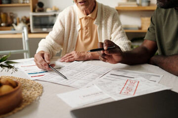 Senior Caucasian woman and Black man reviewing financial documents together at table, discussing bills and payments, hands pointing at paperwork with overdue notice visible