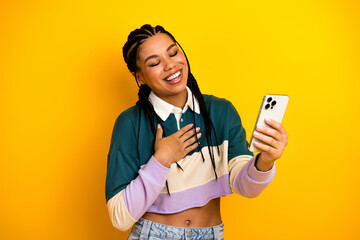 Young woman with braided hair holding a smartphone and smiling happily against a bright yellow...