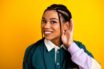 Cute woman with braided hair smiling brightly while holding one hand to ear against yellow...