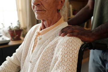 Senior Caucasian woman sitting in wheelchair with disability, Black man standing behind gently placing hand on her shoulder, both partially visible in indoor setting