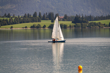 Sailboat gliding across calm lake waters surrounded by lush greenery and mountains in bright...
