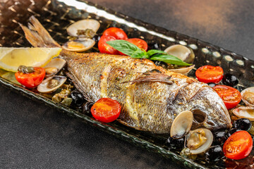 Baked whole fish with tomatoes olives and clams on a black plate. top view. copy space