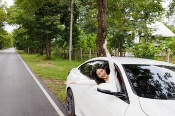 A young Asian cute girl happy smile from a car window sitting in a driver position beside a road while travel in the mountain and tropical forest trees