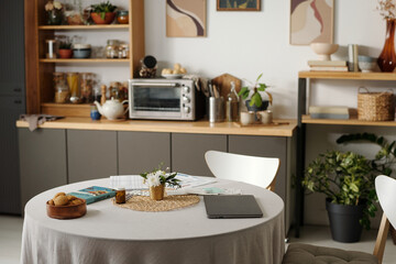 Round table with digital tablet, documents, small vase with flowers and plate of cookies standing in modern kitchen interior with shelves, plants and kitchen appliances visible in background