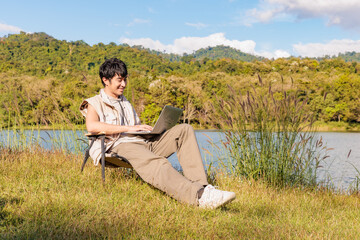 A young Asian man is happy smiling working with his laptop notebook while traveling and camping beside a lake on sunny day
