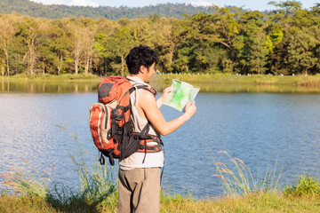 An Asian young man traveler looking at an old paper map to find a route beside a lake during sunny day