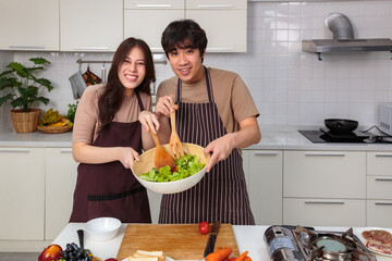 Loving young Asian cooking salad together in home kitchen looking at camera