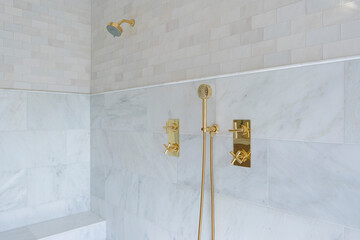 Detail of walk in shower with light colored stone and tile walls, bench seat, and gold hardware.