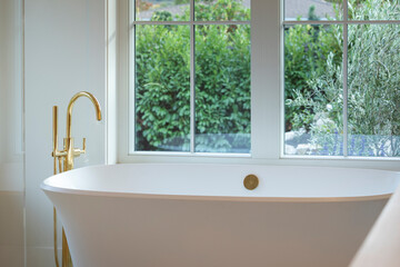 Relaxing scene of soaking tub with gold faucet and view of greenery.