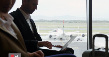 Typing businessman sitting in airport departure lounge, with laptop, suitcase beside large window