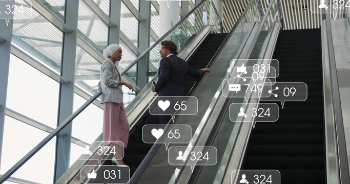 Talking business colleagues climbing escalator holding rail in glass atrium, with social icons - Powered by Adobe
