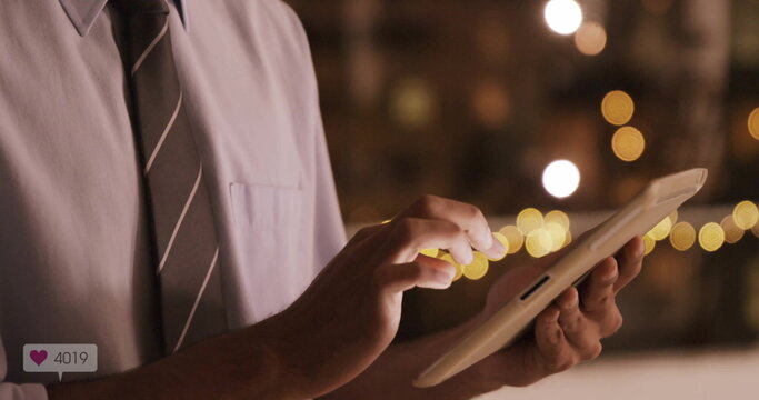 Tapping businessman in shirt and tie holding touchscreen tablet in lounge, with warm string lights