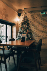 Christmas tree with ornaments standing by a wooden table and chairs inside a cozy decorated cafe