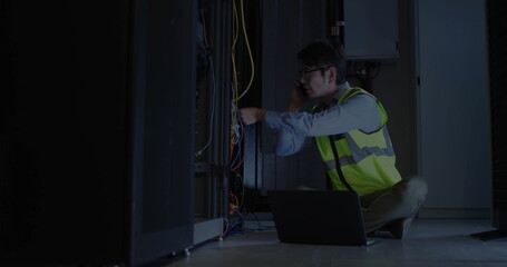 Technician wearing safety vest, glasses connecting cables in server room, with smartphone, laptop
