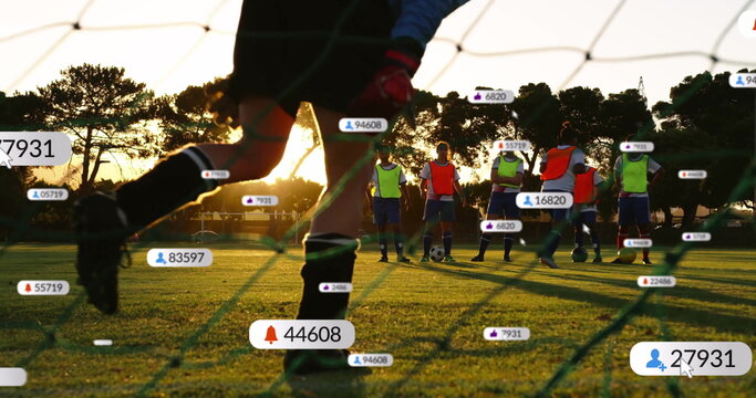 Goalkeeper in blue jersey kicking soccer ball on pitch at sunset, with floating social icons - Powered by Adobe