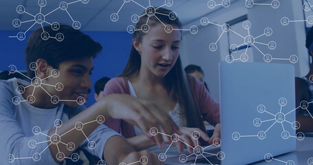 Typing teenage girl and pointing bi-cultural boy at classroom desk, showing laptop network icons