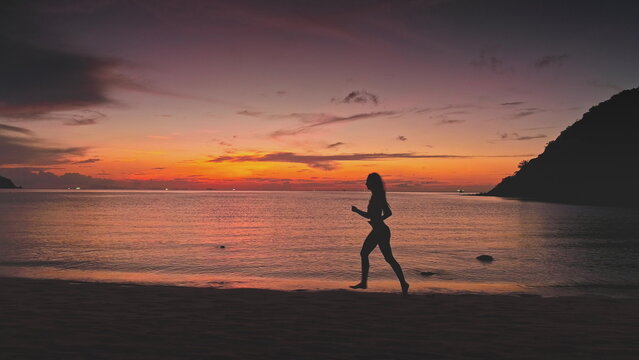 Silhouette of sporty woman running barefoot on the beach in Thailand, enjoying stunning sunset over calm sea with colorful sky and distant islands