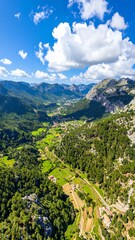 A high-angle, expansive view of a picturesque valley nestled amongst towering mountains, dotted with farms and dwellings, bathed in the bright sunlight of a clear day.