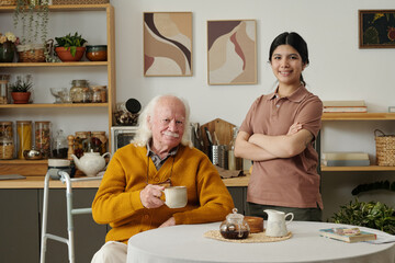 Portrait of senior Caucasian man with long hair holding mug sitting at table with walker beside him, smiling girl with dark hair standing next to him with arms crossed