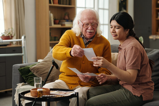 Senior Caucasian man sitting on sofa sharing old photographs with young adult Hispanic woman, both smiling and engaging in conversation, walking cane and walker visible nearby