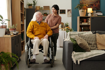Senior Caucasian man with disability sitting in wheelchair talking to young adult Latin woman standing behind him in living room, surrounded by houseplants and cozy decor