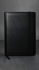 A black leather-like notebook, featuring a simple design and a textured surface, sits on a dark gray surface.