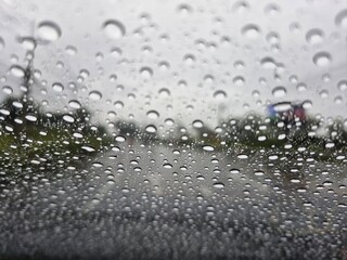 A close-up shot of raindrops on a car windshield on a rainy day. The water droplets vary in size and shape, with slight reflections. The background shows a blurred view of a road and trees, creating a