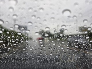 A close-up shot of raindrops on a car windshield on a rainy day. The water droplets vary in size and shape, with slight reflections. The background shows a blurred view of a road and trees, creating a