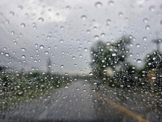 A close-up shot of raindrops on a car windshield on a rainy day. The water droplets vary in size and shape, with slight reflections. The background shows a blurred view of a road and trees, creating a