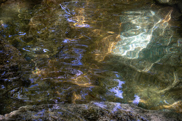 Sunlight reflections on clear water with visible stones at the bottom. Natural abstract background with ripples, light patterns, and colorful texture