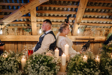 A bride and groom sit back-to-back during their wedding reception, playfully holding up each other’s shoes in a rustic venue decorated with flowers and candles.