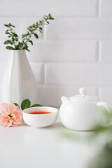 A cup of red rose tea served with a white teapot on a clean white kitchen counter top, complemented by a refreshing view of lush green leaves