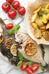 Tasty roasted flounder fish served with potatoes, tomatoes, basil and lemon slices on white marble table, flat lay. Homemade seafood dish