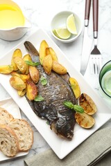 Tasty roasted flounder fish served with potatoes, basil and lemon slices on white marble table, flat lay. Homemade seafood dish