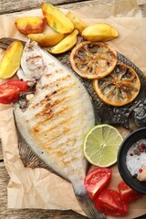 Tasty roasted flounder fish served with potatoes, tomatoes and lemon slices on wooden table, top view. Homemade seafood dish