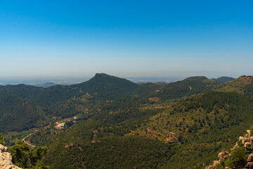 Dramatic rock formations rise above lush greenery at El Garbi, a popular lookout point in the Serra Calderona range of Spain. The panoramic view under a clear blue sky.