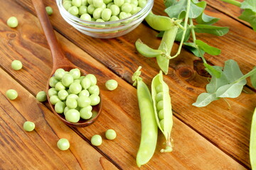There is a glass transparent cup and a spoon with green fresh peas on the wooden table.	