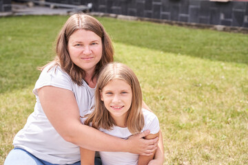 Caucasian adult female and young girl sitting on grass with smile outdoors in sunlit yard.