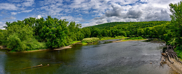 This panoramic image captures a serene river winding through a lush, green landscape under a partly cloudy sky. The far bank is covered with dense foliage, while a forested hill rises in the distance.