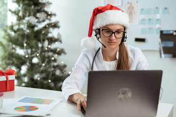 Female doctor in santa hat consulting patient using headset and laptop on shift on christmas eve in...