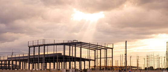 Silhouette metal structure of large factory industrial building framework in construction site against cloudy sky at sunset time, panoramic view