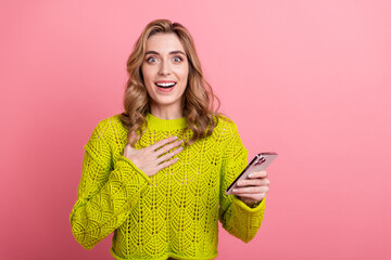 Cheerful young woman holding smartphone in hand against pink background, cheerful expression in...