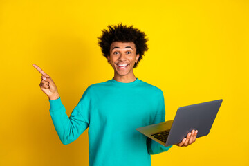 Young man pointing and holding a laptop on a vibrant yellow background exuding excitement and positivity