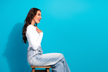 Stylish young woman wearing casual attire sitting on a stool against a vibrant blue background,...
