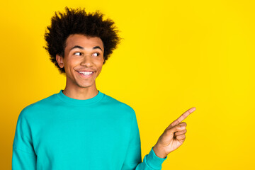 Cheerful young man in a teal sweatshirt pointing to the side against a bright yellow background...