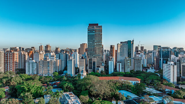 Drone aerial view of S&atilde;o Paulo city skyline along Avenida 9 de Julho with tall modern skyscrapers, residential buildings, green trees and blue clear sky in Brazil