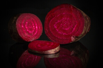 The photo shows a beetroot cut into several pieces on a dark, reflective surface. The rich dark red flesh with characteristic ring patterns is visible.
